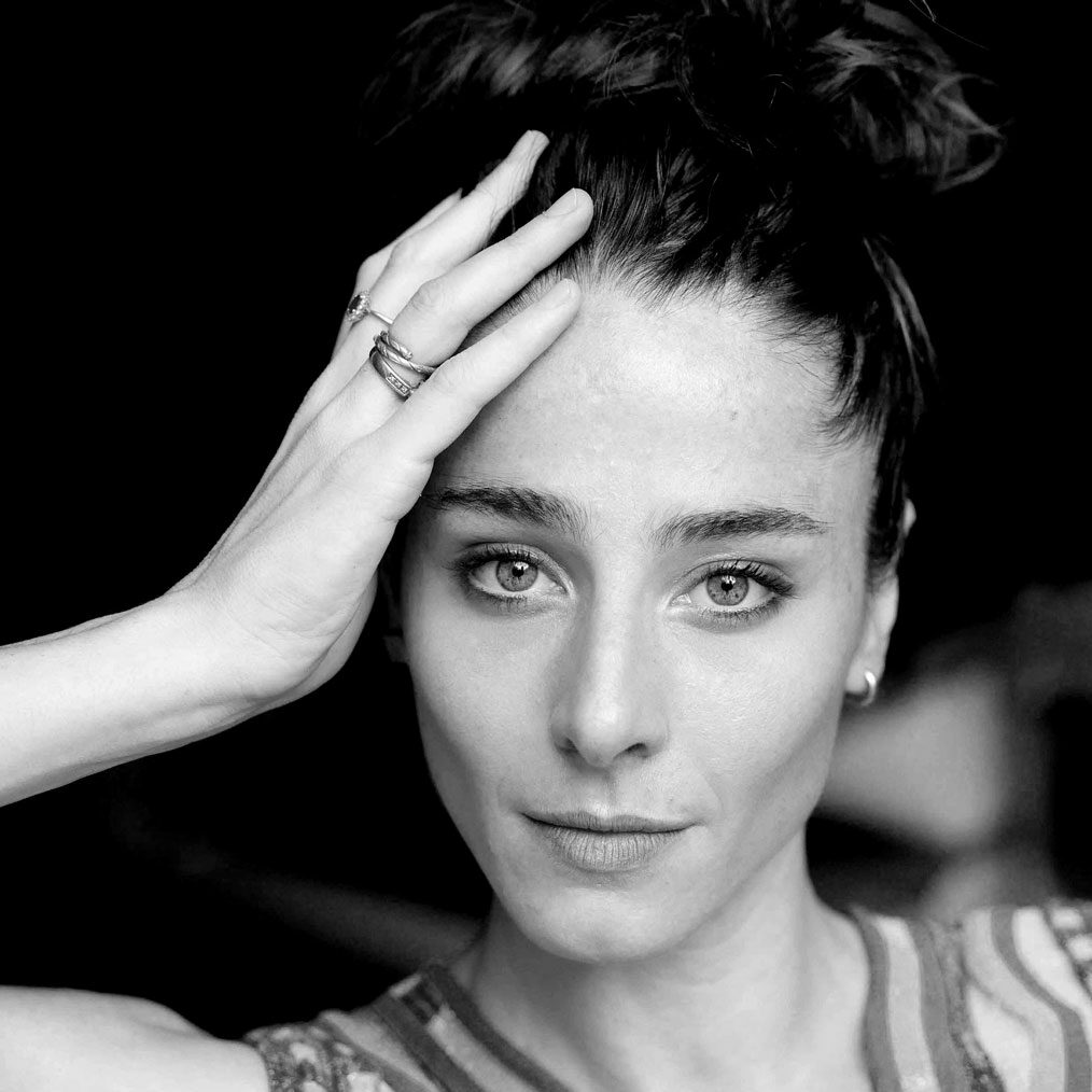 Schwarz-weißes Nahporträt: Caterina Gabanella mit hochgesteckten Haaren legt eine Hand mit Ringen an die Stirn und blickt direkt in die Kamera.